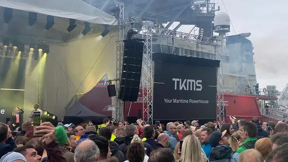 Menschen stehen vor einer Bühne auf dem Werftgelände von Thyssenkrupp in Kiel. Auf einer Leinwand ist das Logo des neuen Markenauftritts als "TKMS" zu sehen.