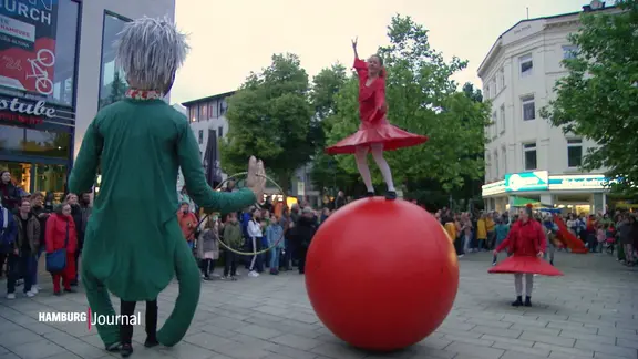 Auf einem öffentlichen Platz schauen viele Menschen einer Tänzerin auf einer großen Kugel und einem Puppenspieler mit einer übergroßen Puppe zu.
