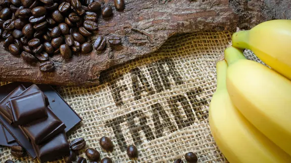 Fair gehandelter Kakao soll Bauern in den Herkunftsländern eine menschenwürdige Existenz sichern. | fotolia.com, Visions-AD Auf einem Sack mit der Aufschrift "Fair Trade" liegen Kaffeebohnen, Bananen und Schokolade.