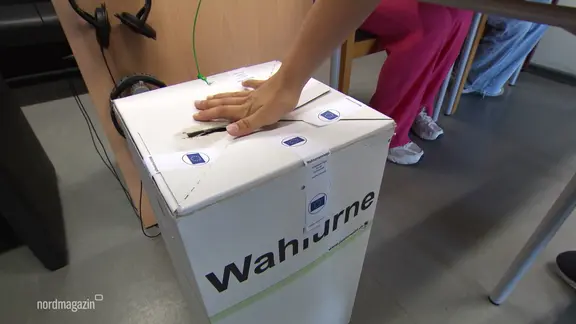 Eine Hand schiebt einen Zettel in eine Wahlurne.
