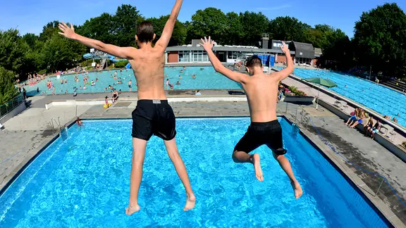 Zwei Jungen springen von einem hohen Sprungturm in ein Schwimmbecken in einer gut besuchten Badeanstalt.