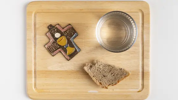 Holzbrett mit einem Kreuz, einem Glas Wasser und einem Stück Brot