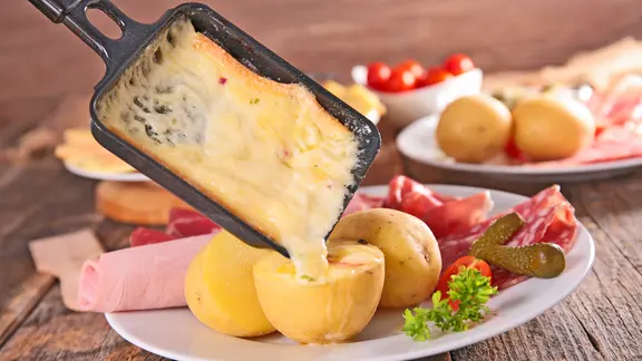 Geschmolzener Raclette-Käse fließt aus einem Pfännchen auf einen Teller mit Kartoffeln und Wurst | M.studio - Fotolia Geschmolzener Raclette-Käse fließt aus einem Pfännchen auf einen Teller mit Kartoffeln und Wurst