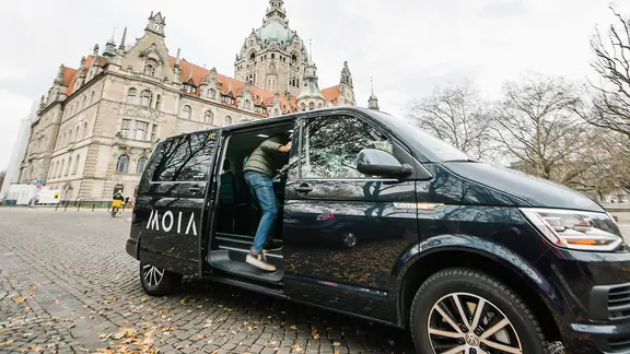 Fahrzeug des Moia-Sammeltaxi am Trammplatz in Hannover mit dem Rathaus im Hintergrund, Fahrgast steigt ein.