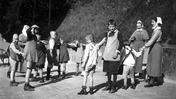 Geistig behinderte Kinder spielen mit zwei Diakonieschwestern um 1930 Ringelreihen.