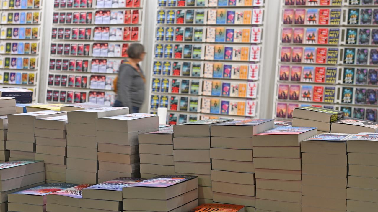 Viele Taschenbücher liegen auf Bücherstapeln bei der Frankurter Buchmesse - im Hintergrund steht eine Frau vor den bunten Covern