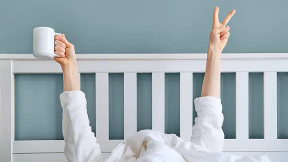 Eine Person, die sich unter einer Decke versteckt, zeigt seine Hand mit dem Siegeszeichen und hält eine Tasse Kaffee.