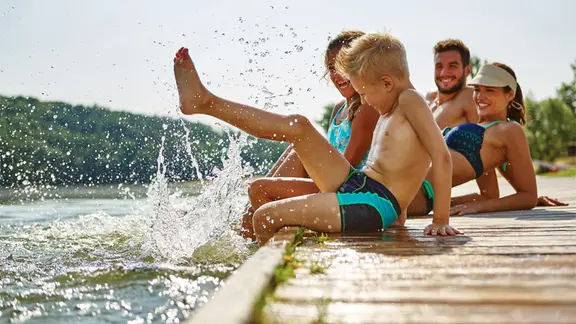Eine Familie sitzt in ausgelassener Stimmung auf einem Steg am Badesee und hat die Füße im Wasser.