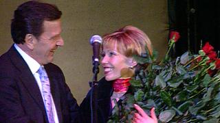 Doris Schröder-Köpf und Gerhard Schröder stehen mit einem Blumenstrauß vor einem Mikrofon.
