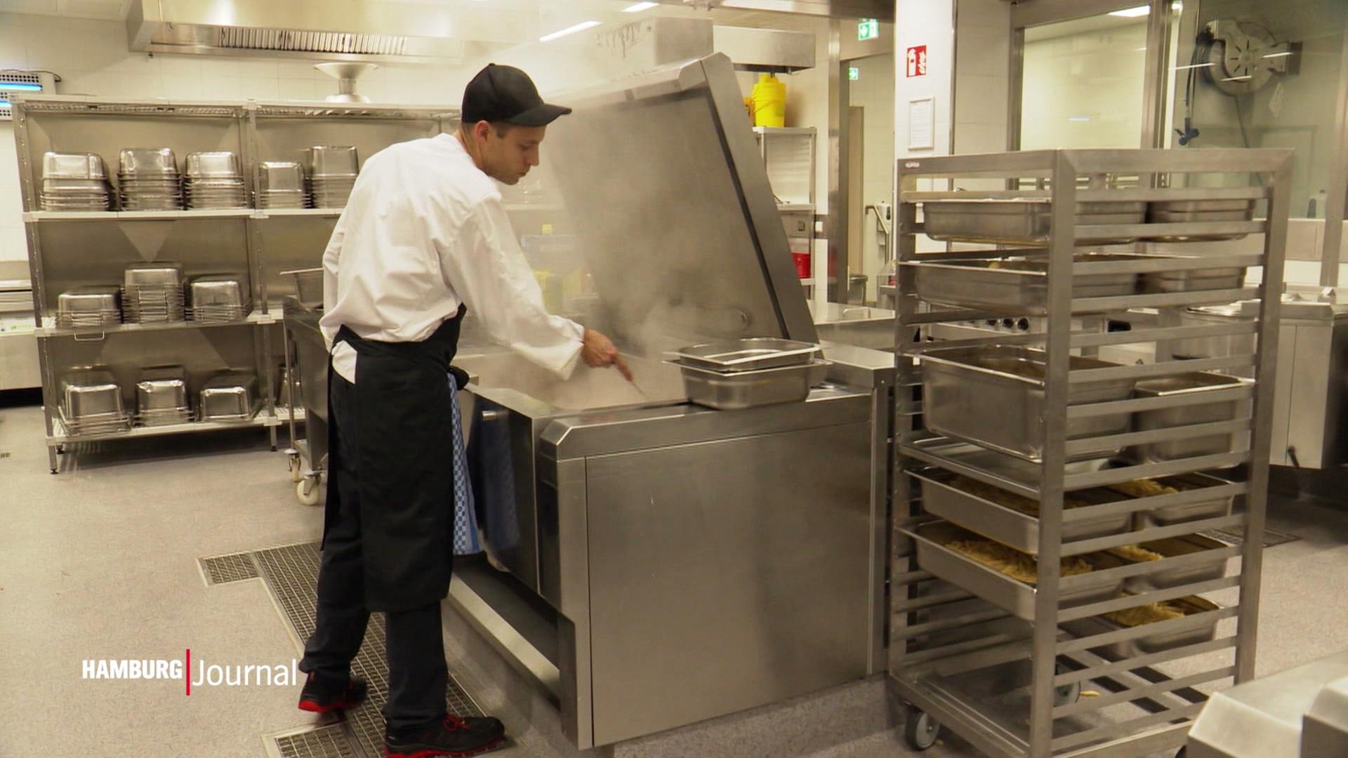 Ein Koch steht in einer Gastroküche und rührt in einem großen Edelstahl-Gerät, aus dem es dampft. | Screenshot