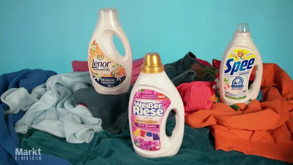 Verschiedene Flüssigwaschmittel Produkte stehen auf einem Tisch umgeben von Klamotten. | Screenshot Verschiedene Flüssigwaschmittel Produkte stehen auf einem Tisch umgeben von Klamotten.