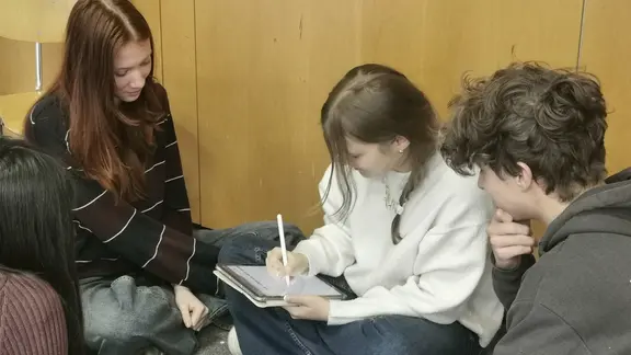 Ein paar Jugendliche sitzen zusammen. Sie sind über ein Tablet gebeugt, auf dem sie etwas schreiben.
