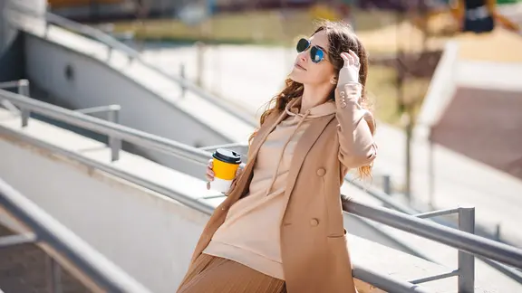 Eine Frau steht mit einem Kaffeebecher an einem Geländer in der Sonne.