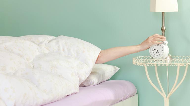 Person greift aus dem Bett nach einem Wecker.