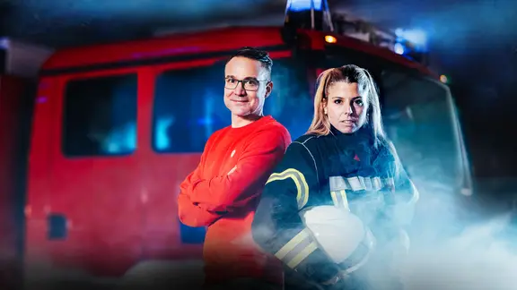 Ein Mann in rotem Pullover und eine Frau in Feuerwehruniform mit Helm stehen vor einem roten Feuerwehrauto mit Blaulicht und Rauch im Hintergrund.