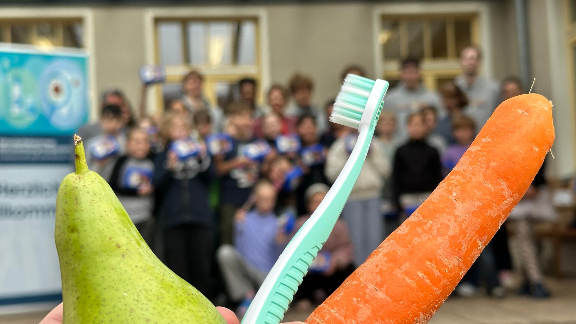Karotte, Zahnbürste, Apfel hochgehalten vor einer Schulklasse im Hintergrund | Denis Mollenhauer