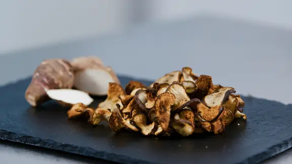 Eine angeschnittene Topinambur-Knolle sowie hauchdünne Topinambur-Chips liegen auf einer Schieferplatte.