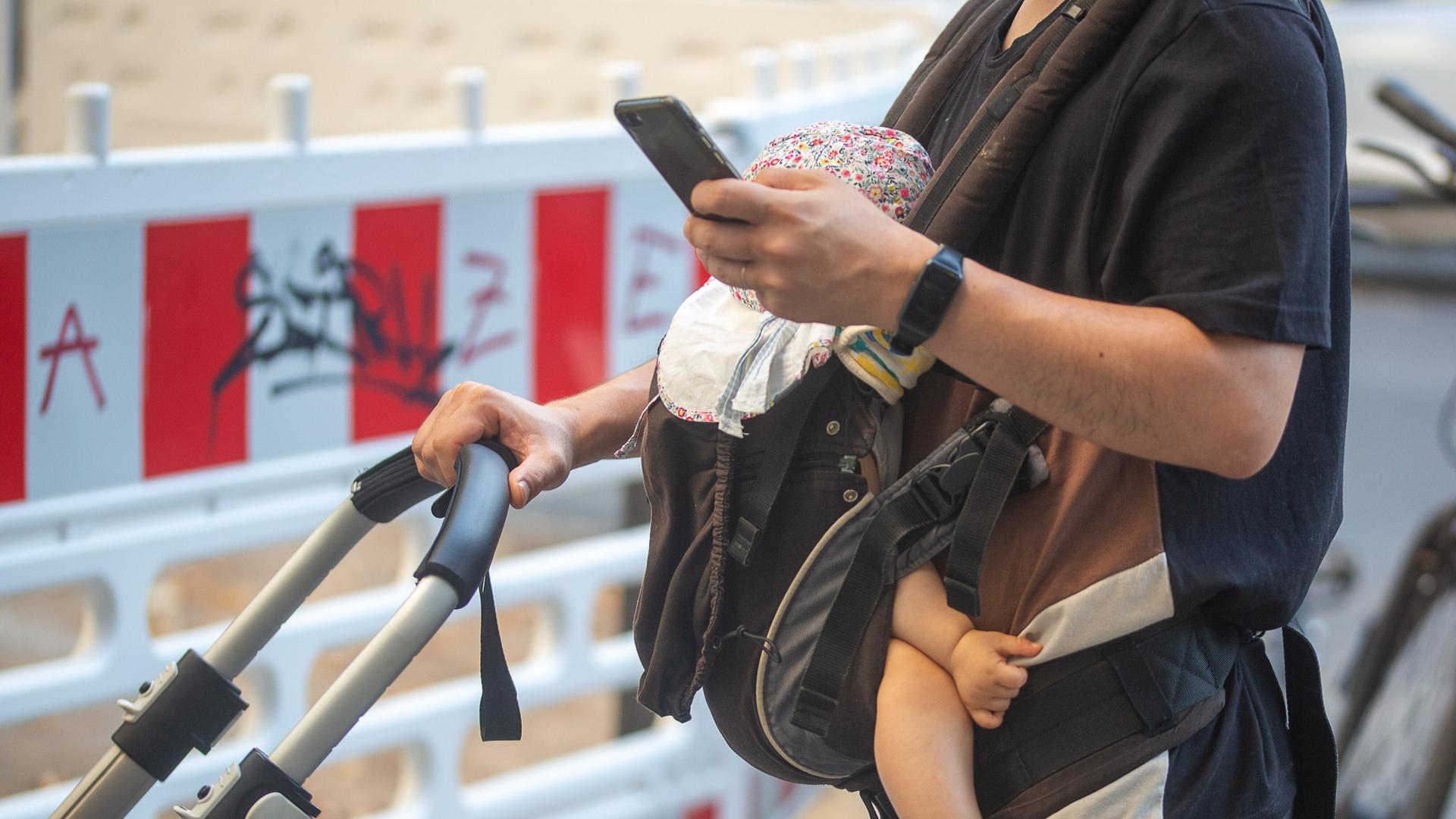 Ein Mann schiebt einen Kinderwagen und trägt gleichzeitig ein Baby in einer Tragehilfe. Dabei schaut er auf sein Smartphone. | picture alliance/dpa | Fernando Gutierrez-Juarez