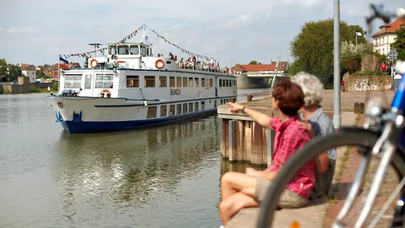 Touristen sitzen am Ufer der Weser in Hameln und schauen auf ein vorbeifahrendes Weserschiff.