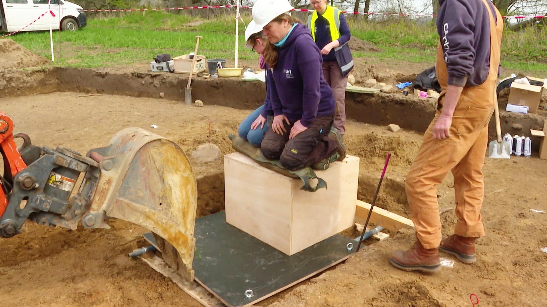 Eine Frau sitzt auf einer Holzkiste mit einem archäologischen Fund. | Screenshot