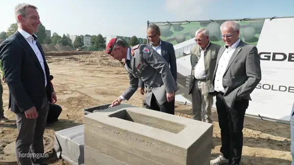 Ein Mann in Militäruniform und weitere Männer in Anzügen legen einen Grundstein für ein neues Gebäude.