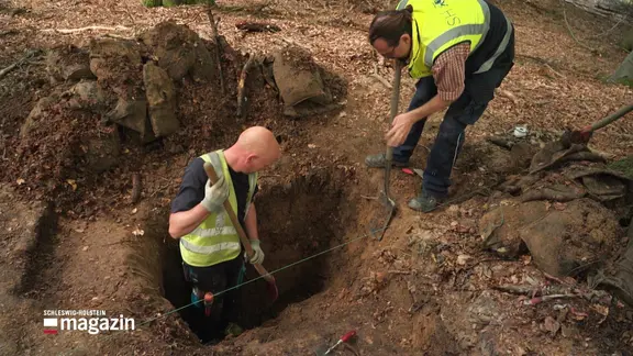 Zwei Männer mit Schaufeln graben ein Loch im Wald.