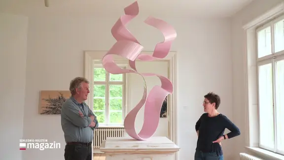 Elke Scheigart und Volker Tiemann stehen einander gegenüber. Zwischen ihnen steht eine Skulptur, die aussieht, als würde sie ein sich bewegendes Geschenkband sein.