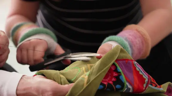 Drei Hände halten Stoff und eine Schere, mit der gerade ein Faden abgeschnitten wird.