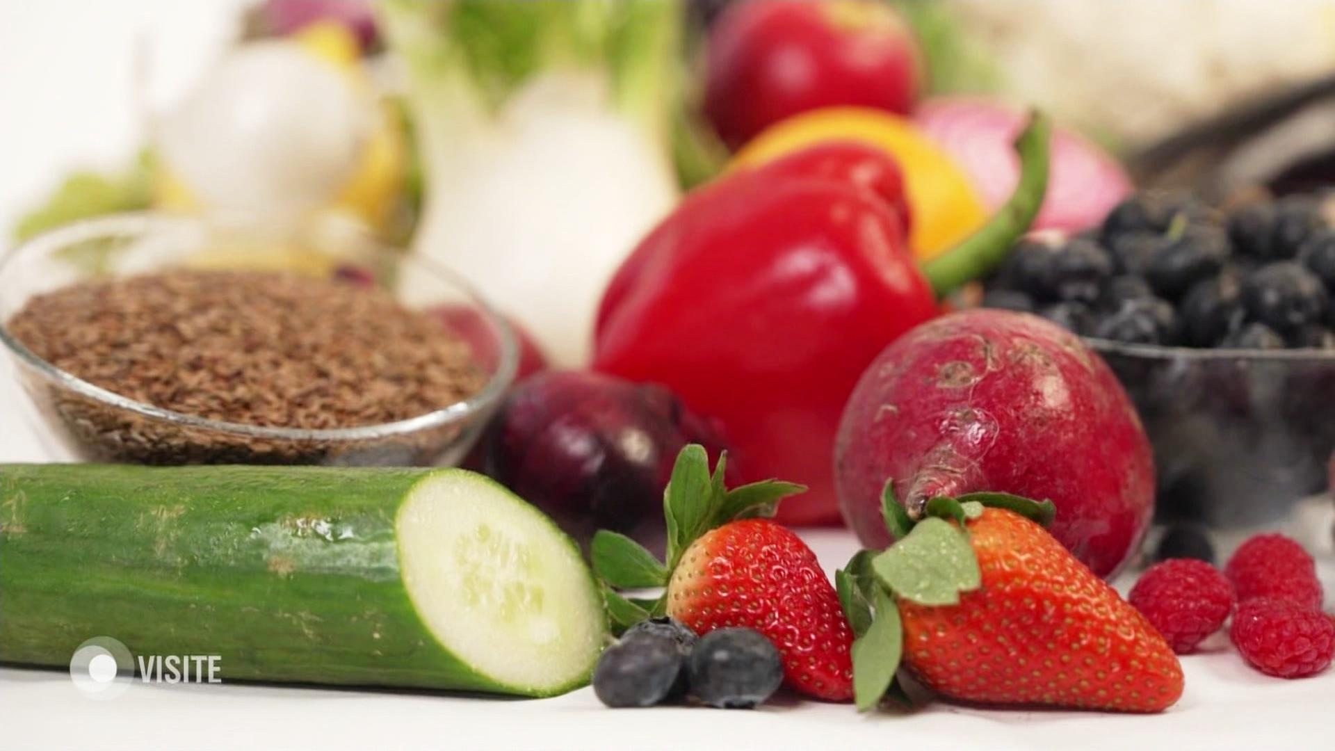 Eine angeschnittene Salatgurke, Obst wie Blaubeeren und Erdbeere, im Hintergrund Radieschen, rote Zwiebeln, Paprika und eine Schale mit Leinsamen. | Screenshot