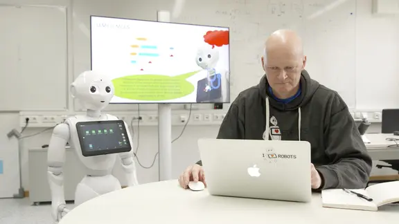 Der KI-Experte Thomas Sievers sitzt am Laptop, neben ihm steht der Roboter Pepper.