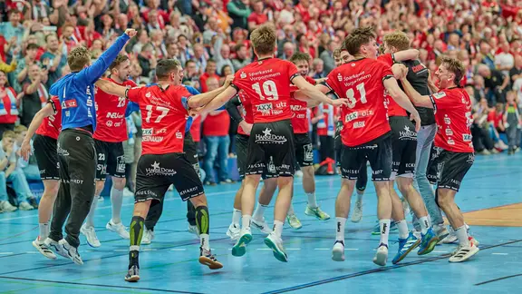 Handballer in roten Trikots jubeln, hüpfen und feiern ihren Sieg vor den Zuschauern in der Halle.
