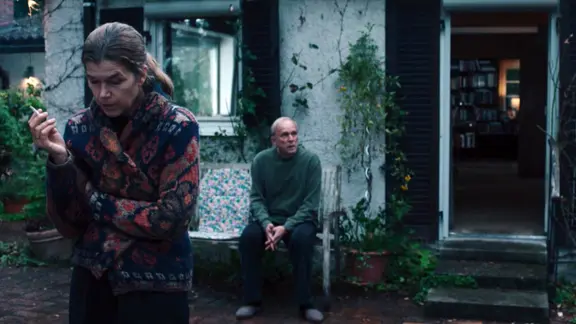 Rita (Anna Engelke) steht und raucht, während Hans (Ulrich Tukur) im Hintergrund auf einer Bank sitzt. 