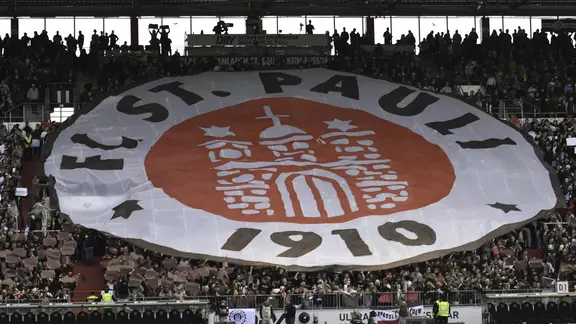 Ein überdimensionales FC St. Pauli-Logo wird von Zuschauerinnen und Zuschauern hochgehalten.