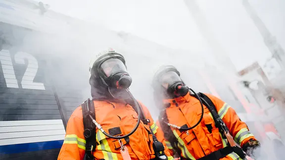 Zwei Feuerwehrleute mit Atemschutzmasken stehen in dichtem Rauch.
