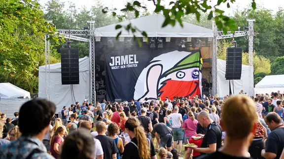 Der Schriftzug "Jamel rockt den Förster" hängt bei einer Umbaupause vor der Bühne. Davor stehen Hunderte Zuschauer.