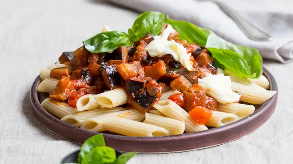 Ein Teller Penne mit Auberginen und Mozzarella steht auf einem Tisch.