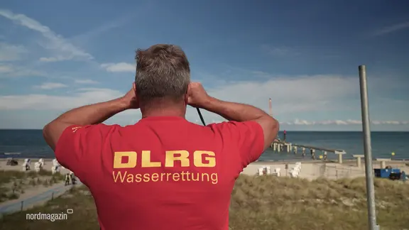 Den beiden Schwestern geht es inzwischen wieder gut. (Themenbild) | NDR Ein Mann mit einem roten DLRG-Shirt schaut mit einem Fernglas aufs Wasser.