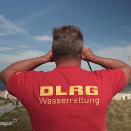 Ein Mann mit einem roten DLRG-Shirt schaut mit einem Fernglas aufs Wasser.