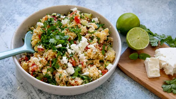 Eine Schüssel Hirse-Tabouleh, daneben Limetten, Kräuter und etwas Feta.