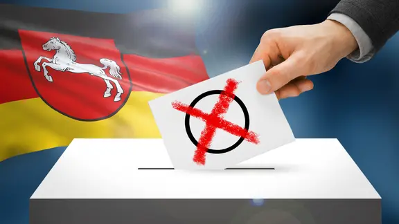 Wahlurne mit Wahlzettel, im Hintergrund das Landeswappen von Niedersachsen