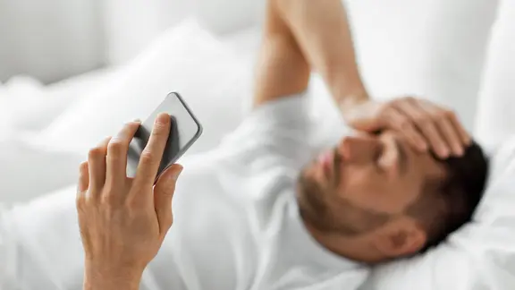 Ein im Bett liegender Mann hält sich mit einer Hand den Kopf, in der anderen ein Smartphone | colourbox Ein im Bett liegender Mann hält sich mit einer Hand den Kopf, in der anderen ein Smartphone