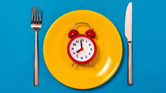 Ein Wecker liegt auf einem gelben Teller, daneben Messer und Gabel auf blauem Hintergrund.