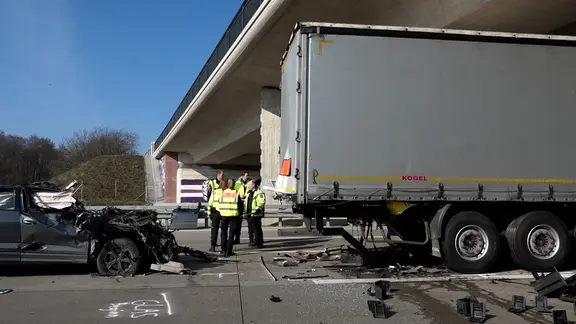 Nach einem tödlichen Unfall auf der A1 bei Holdorf steht ein stark beschädigtes Auto hinter einem Lkw, Polizeikräfte stehen daneben.