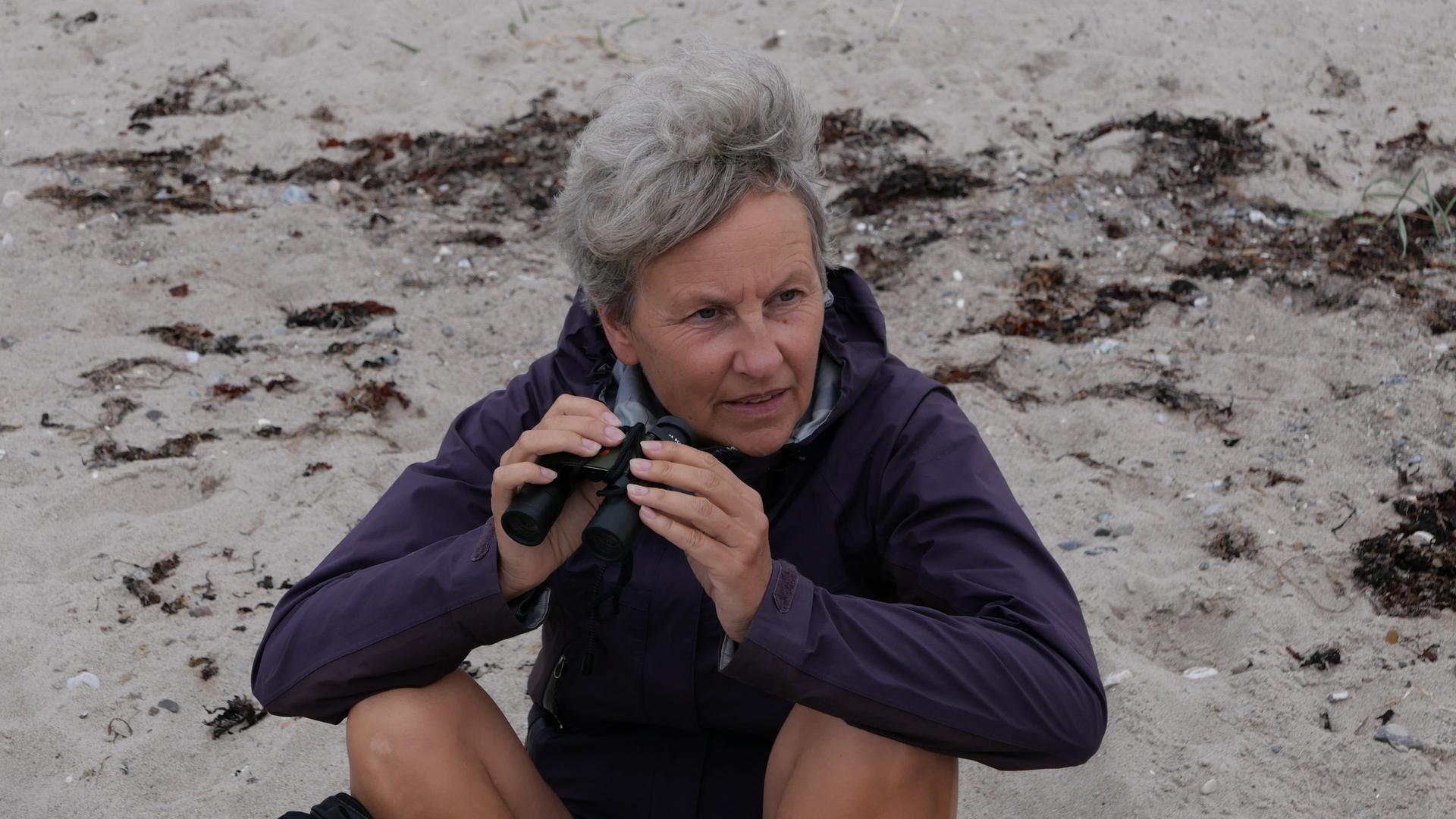 Anja Liedtke sitzt auf Sandboden und hält ein kleines Fernglas in den Händen.  | Anja Liedtke