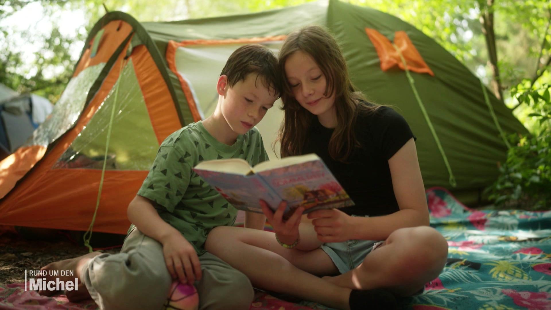Bruder und Schwester lesen gemeinsam ein Buch vor ihren Zelten im Elbecamp. | Screenshot