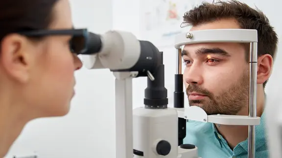 Mann sitzt vor Untersuchungsgerät bei Augenärztin. Eine Lampe beleuchtet seine Iris (Bild: colourbox.de/Syda Productions)
