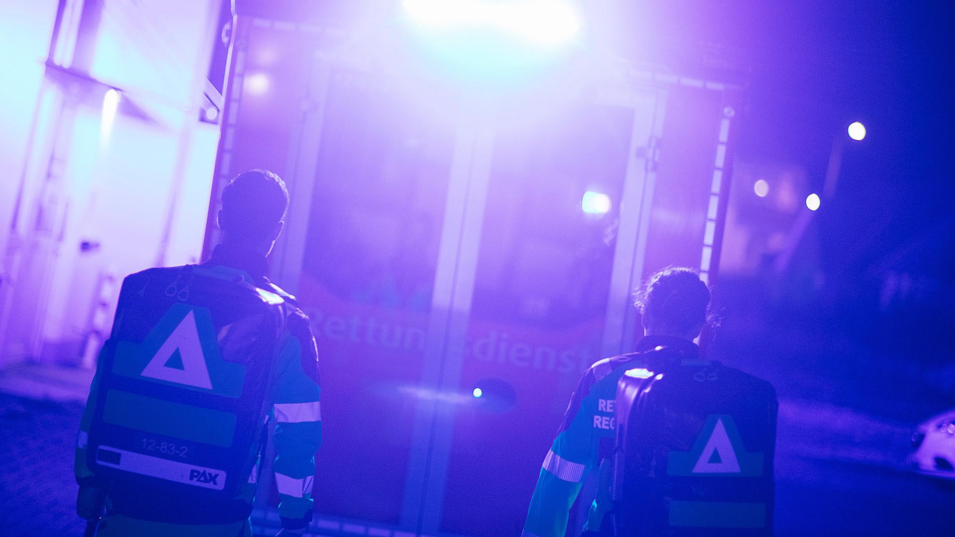 Zwei Sanitäter laufen in Richtung eines Rettungswagens, der sie mit Blaulicht anstrahlt. | NDR, Julius Matuschik