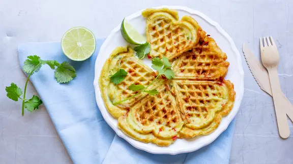 Ein Teller mit einer Avocadowaffel, Limetten und ein Zweig Koriander liegen auf dem Tisch.