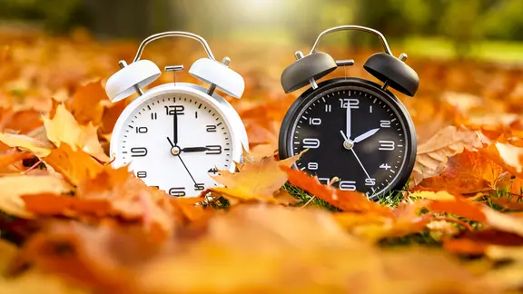 Zwei Wecker im Herbstlaub - einer mit Sommer- einer mit Winterzeit | picture alliance / CHROMORANGE Zwei Wecker im Herbstlaub - einer mit Sommer- einer mit Winterzeit