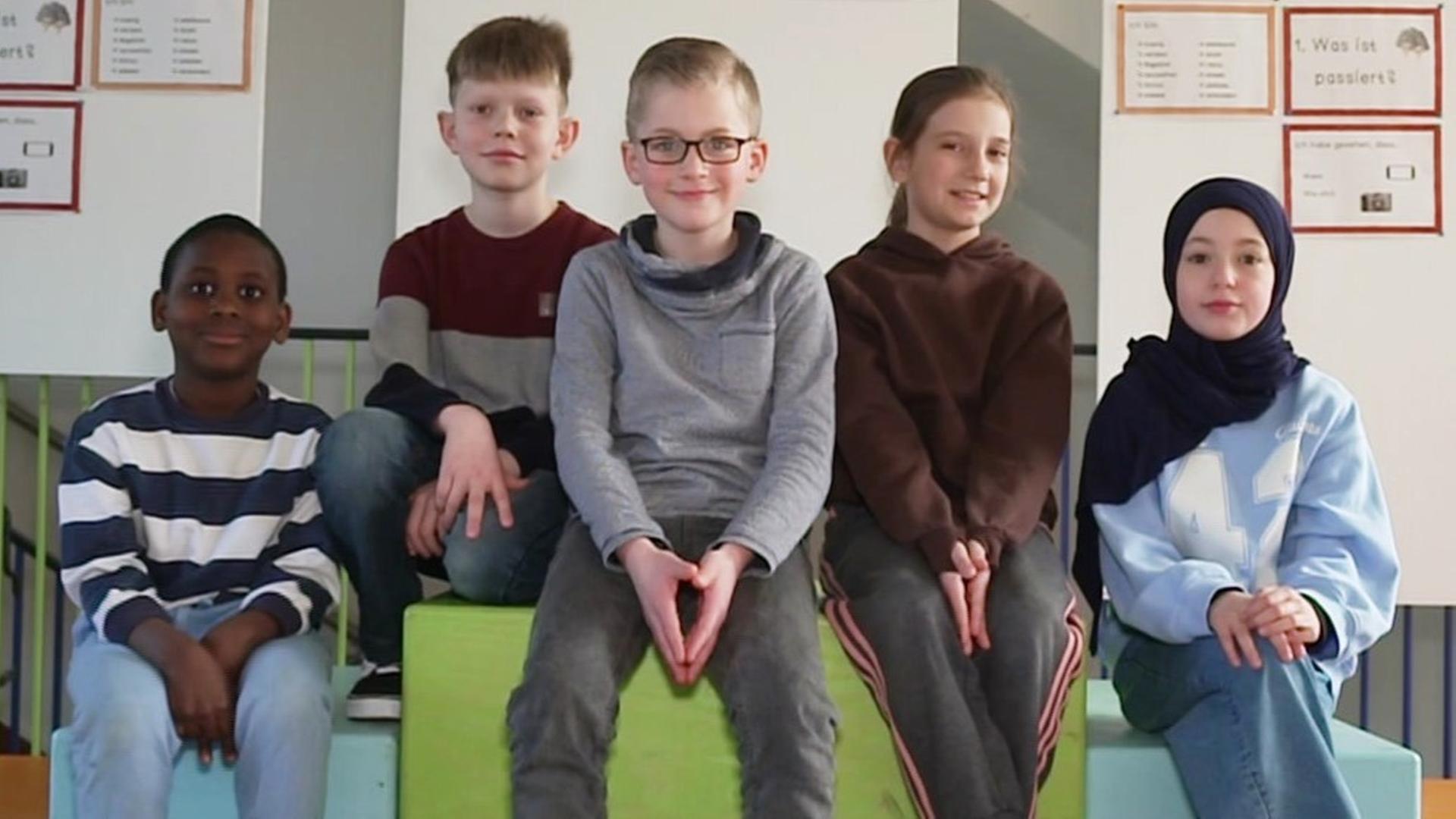 Fünf Grundschulkinder sitzen auf den bunten Stufen einer Friedensbrücke. | Screenshot